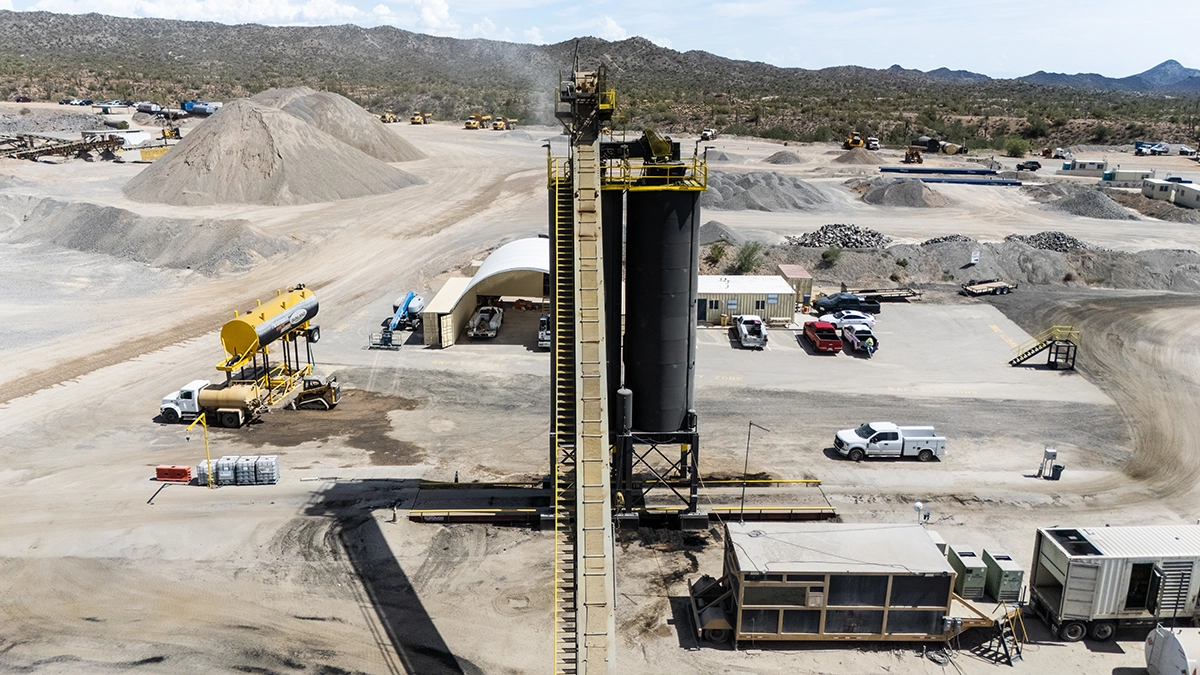 A tall industrial structure stands in a mining or quarry site, surrounded by trucks, equipment, and scattered piles of dirt and gravel under a partly cloudy sky.